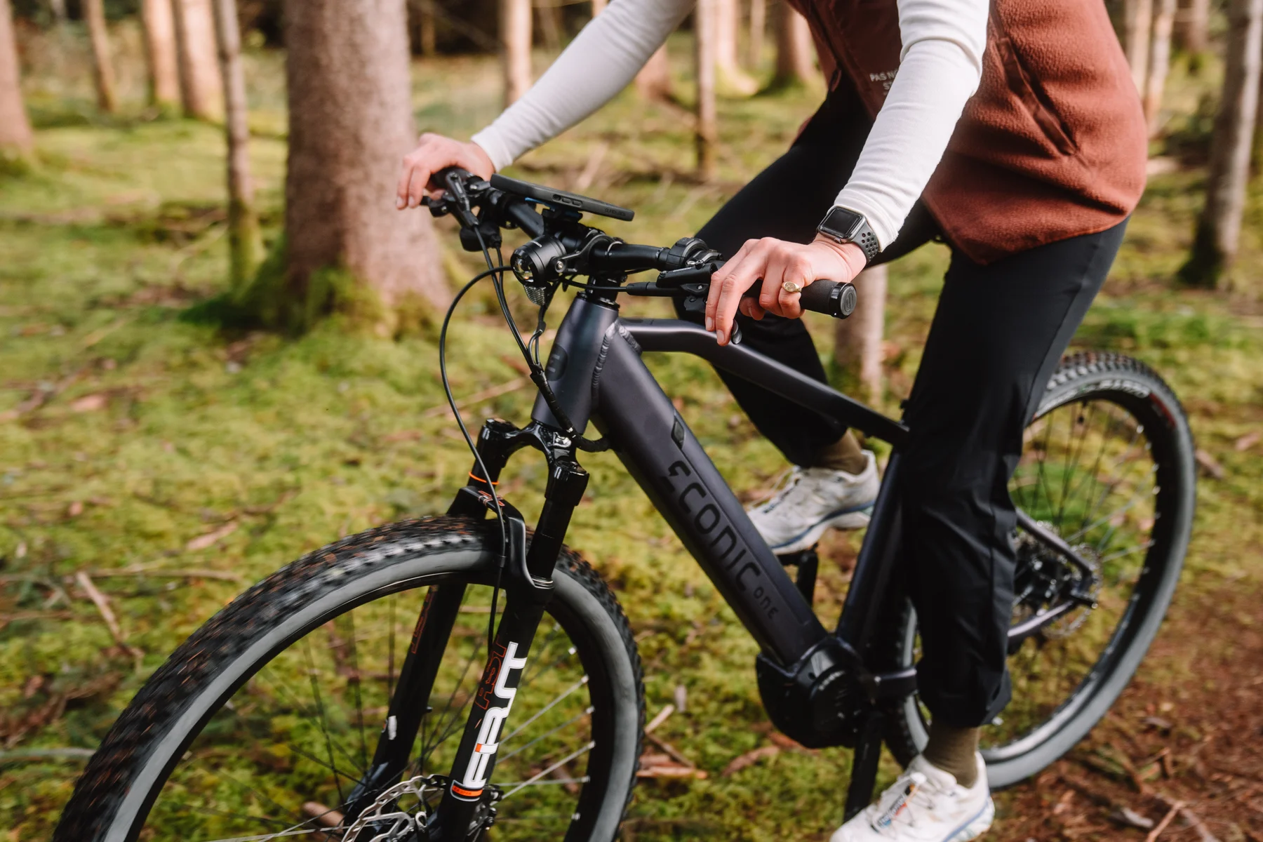 Eonic One Adventure kaufen: Dunkelgraues E-Mountainbike mit Federgabel im echten Offroad-Einsatz auf moosigem Waldboden mit Fahrer.