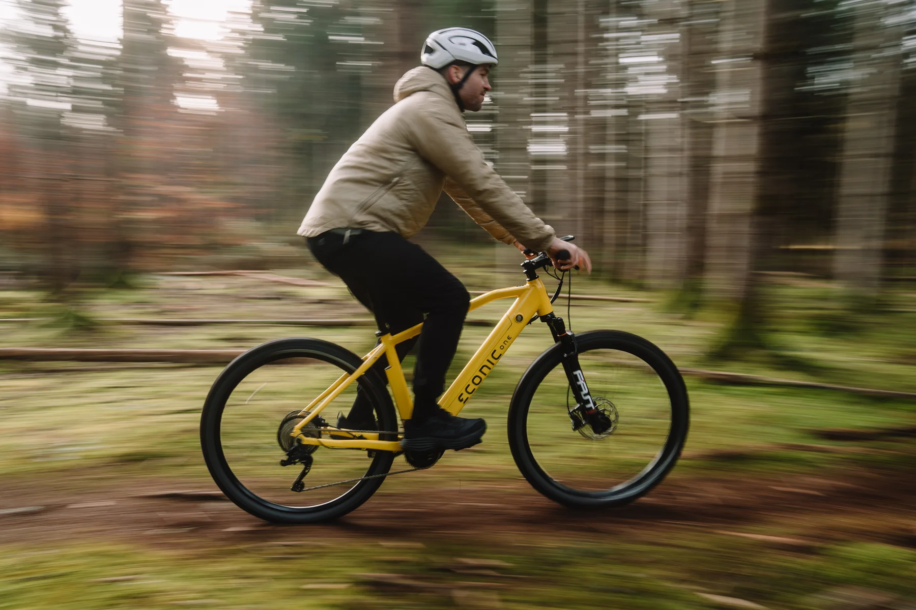 Eonic One Adventure kaufen: Gelbes E-Mountainbike in schneller Fahrt mit einem Radfahrer auf einem kurvigen Trail im dichten Wald.