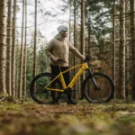 Eonic One Adventure kaufen: Gelbes E-Mountainbike mit einem Radfahrer bei einer Pause auf moosigem Boden in einem dichten Nadelwald.