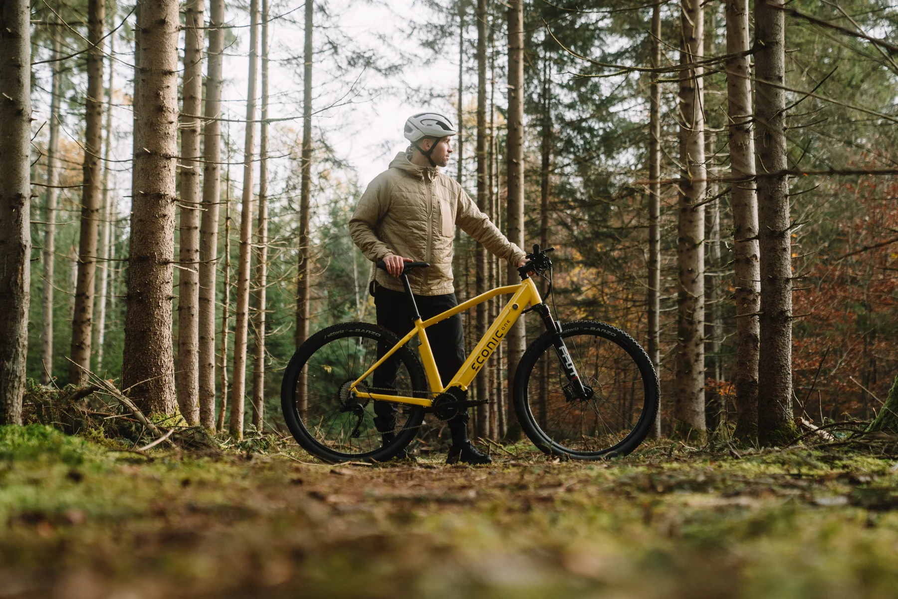 Eonic One Adventure kaufen: Gelbes E-Mountainbike mit einem Radfahrer bei einer Pause auf moosigem Boden in einem dichten Nadelwald.