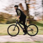Eonic One Bandit kaufen: Sportliches dunkelgraues E-Bike im dynamischen Fahrbetrieb mit einem Radfahrer in einer herbstlichen Parkanlage.