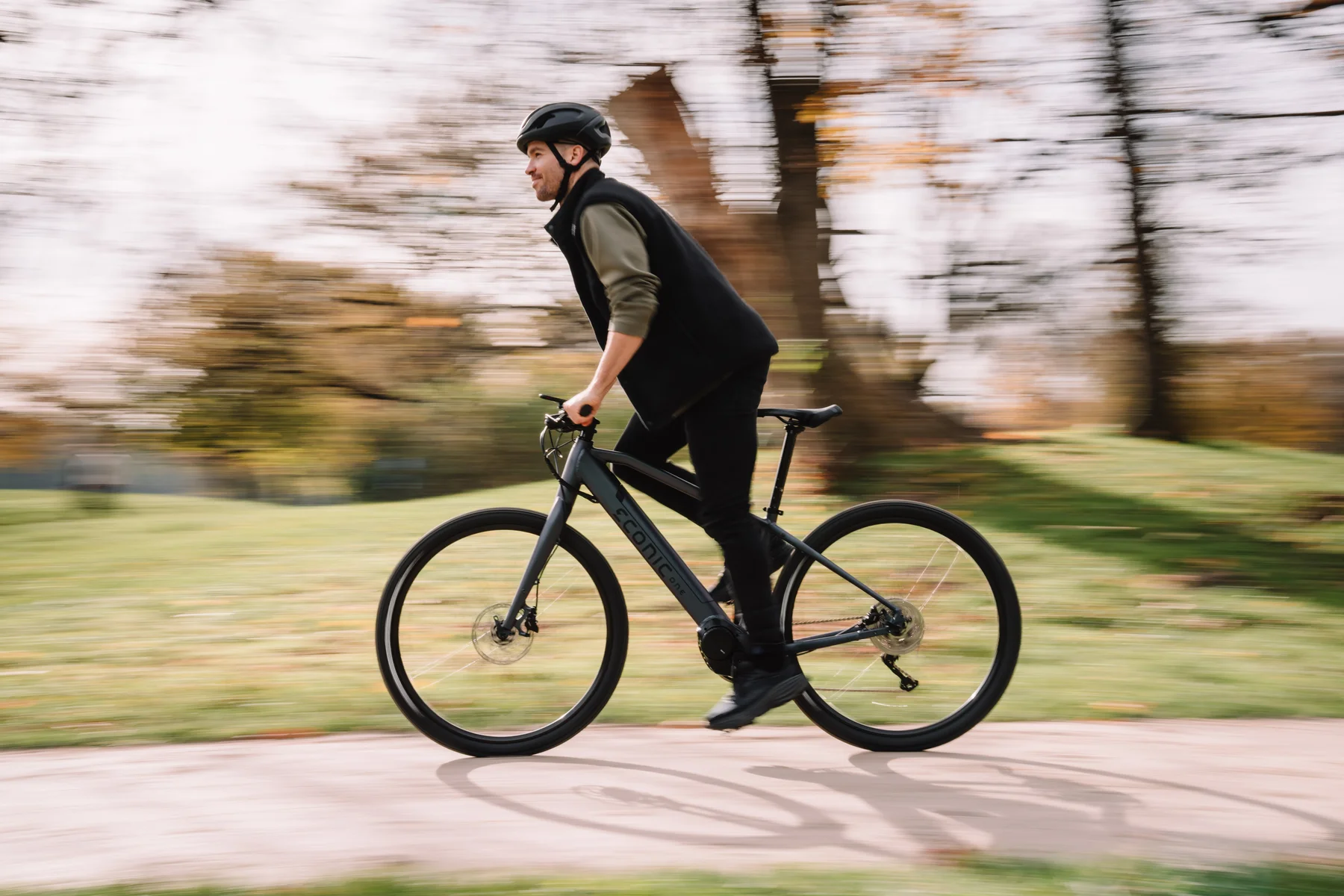Eonic One Bandit kaufen: Sportliches dunkelgraues E-Bike im dynamischen Fahrbetrieb mit einem Radfahrer in einer herbstlichen Parkanlage.