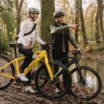 Eonic One Bandit kaufen: Ein dunkelgraues Urban-E-Bike und ein gelbes Modell bei einem gemeinsamen Ausflug eines Paares an einem Fluss im herbstlichen Wald.