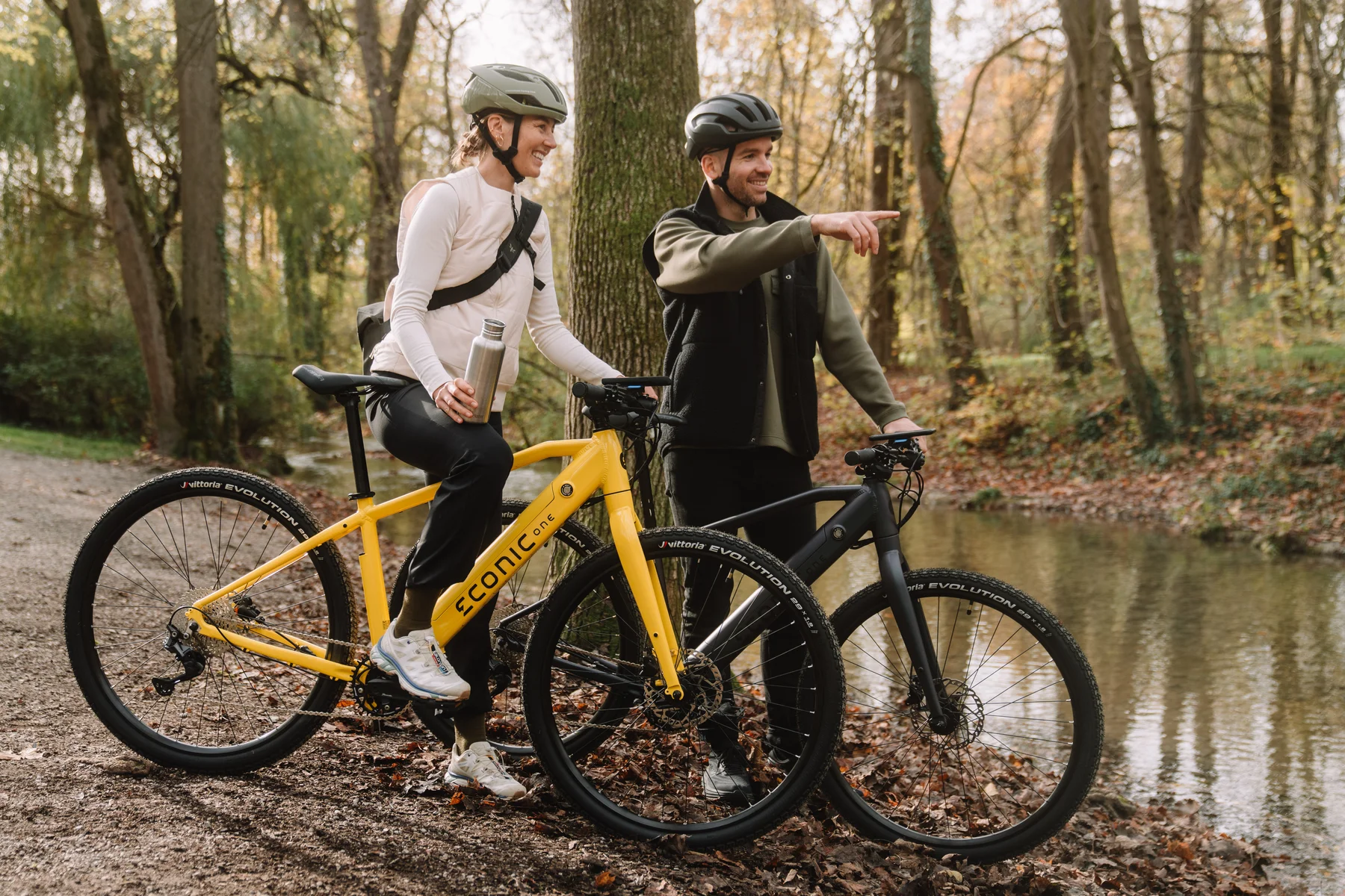 Eonic One Bandit kaufen: Ein dunkelgraues Urban-E-Bike und ein gelbes Modell bei einem gemeinsamen Ausflug eines Paares an einem Fluss im herbstlichen Wald.