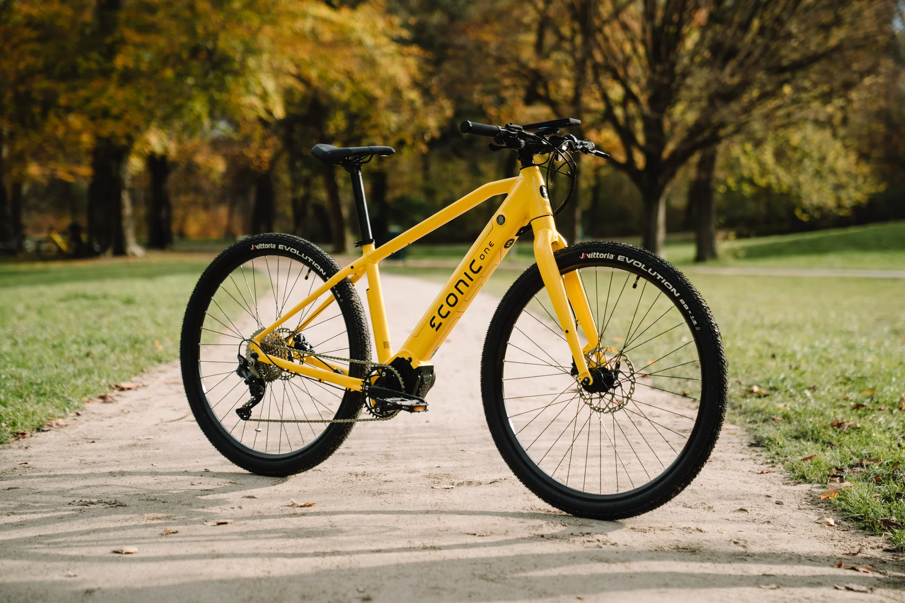 Eonic One Bandit kaufen: Leuchtend gelbes sportliches E-Bike auf einem sandigen Parkweg vor einer herbstlichen Baum- und Wiesenlandschaft.