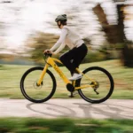 Eonic One Bandit kaufen: Gelbes sportliches E-Bike im schnellen Fahrbetrieb mit einer Radfahrerin in einer herbstlichen Parkanlage.