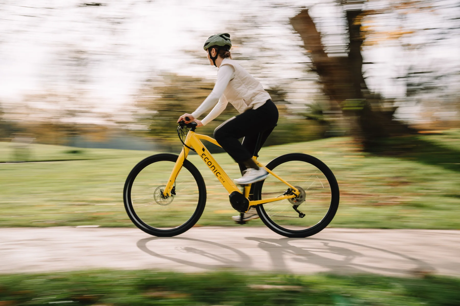 Eonic One Bandit kaufen: Gelbes sportliches E-Bike im schnellen Fahrbetrieb mit einer Radfahrerin in einer herbstlichen Parkanlage.