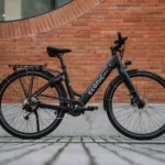 Eonic One Comfort Limited kaufen: Matt-schwarzes E-Bike mit tiefem Einstieg auf Pflastersteinen vor einer modernen rötlichen Backsteinwand.