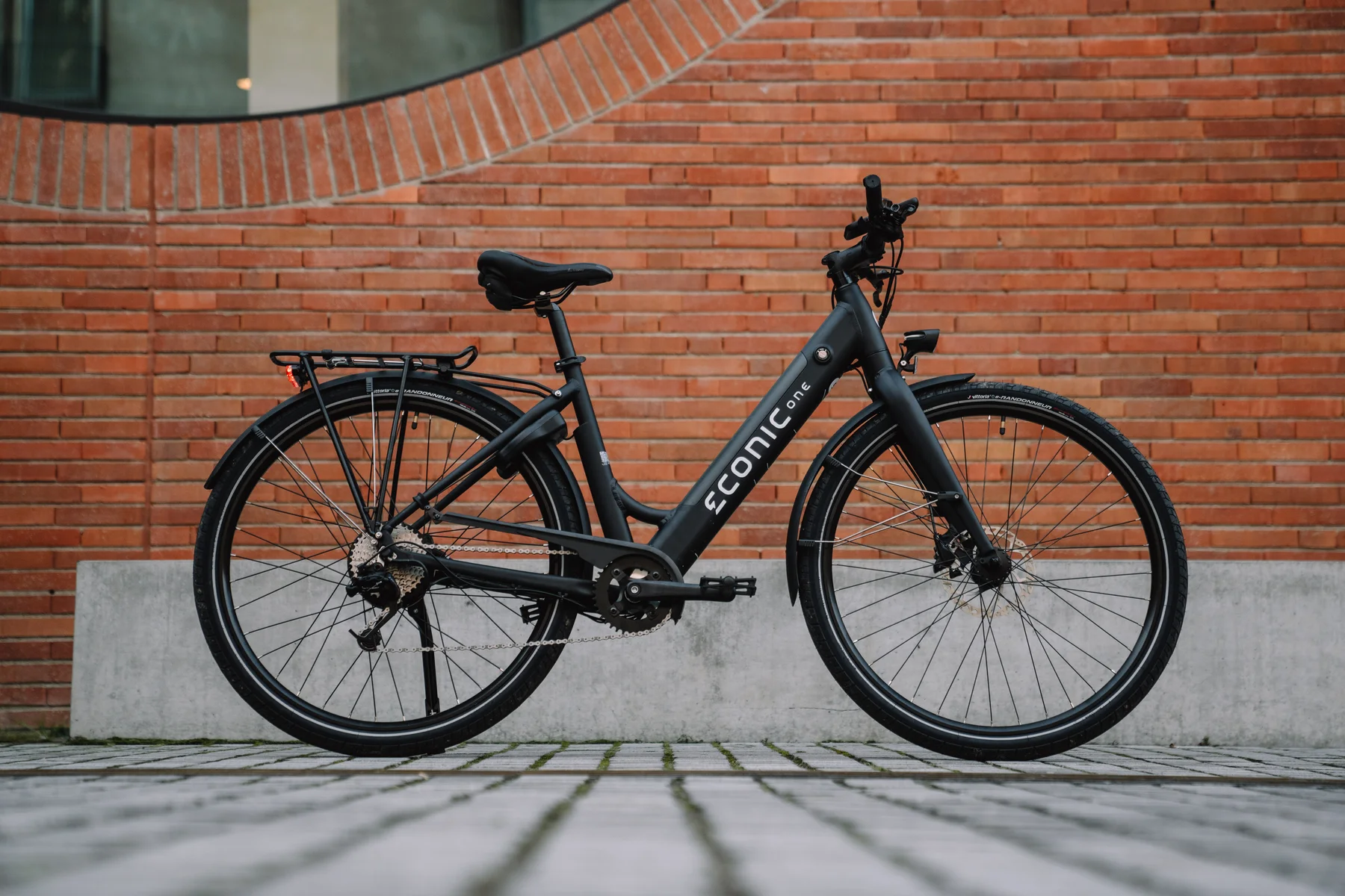 Eonic One Comfort Limited kaufen: Matt-schwarzes E-Bike mit tiefem Einstieg auf Pflastersteinen vor einer modernen rötlichen Backsteinwand.