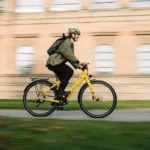 Eonic One Comfort Limited kaufen: Gelbes City-E-Bike im dynamischen Fahrbetrieb mit einem Radfahrer vor einem historischen Gebäude.