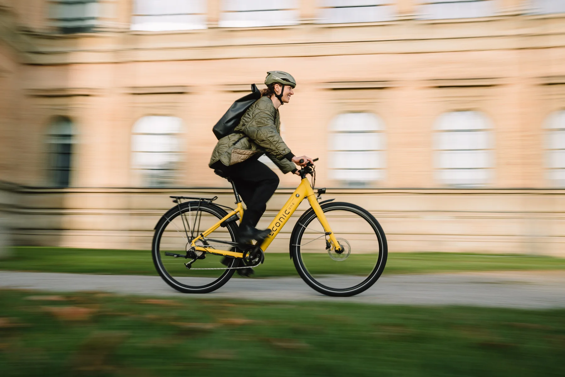 Eonic One Comfort Limited kaufen: Gelbes City-E-Bike im dynamischen Fahrbetrieb mit einem Radfahrer vor einem historischen Gebäude.