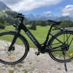Eonic One Comfort Limited kaufen: Matt-schwarzes E-Bike auf einem Schotterweg vor einem beeindruckenden Alpen-Panorama mit grünen Wiesen und Bergen.