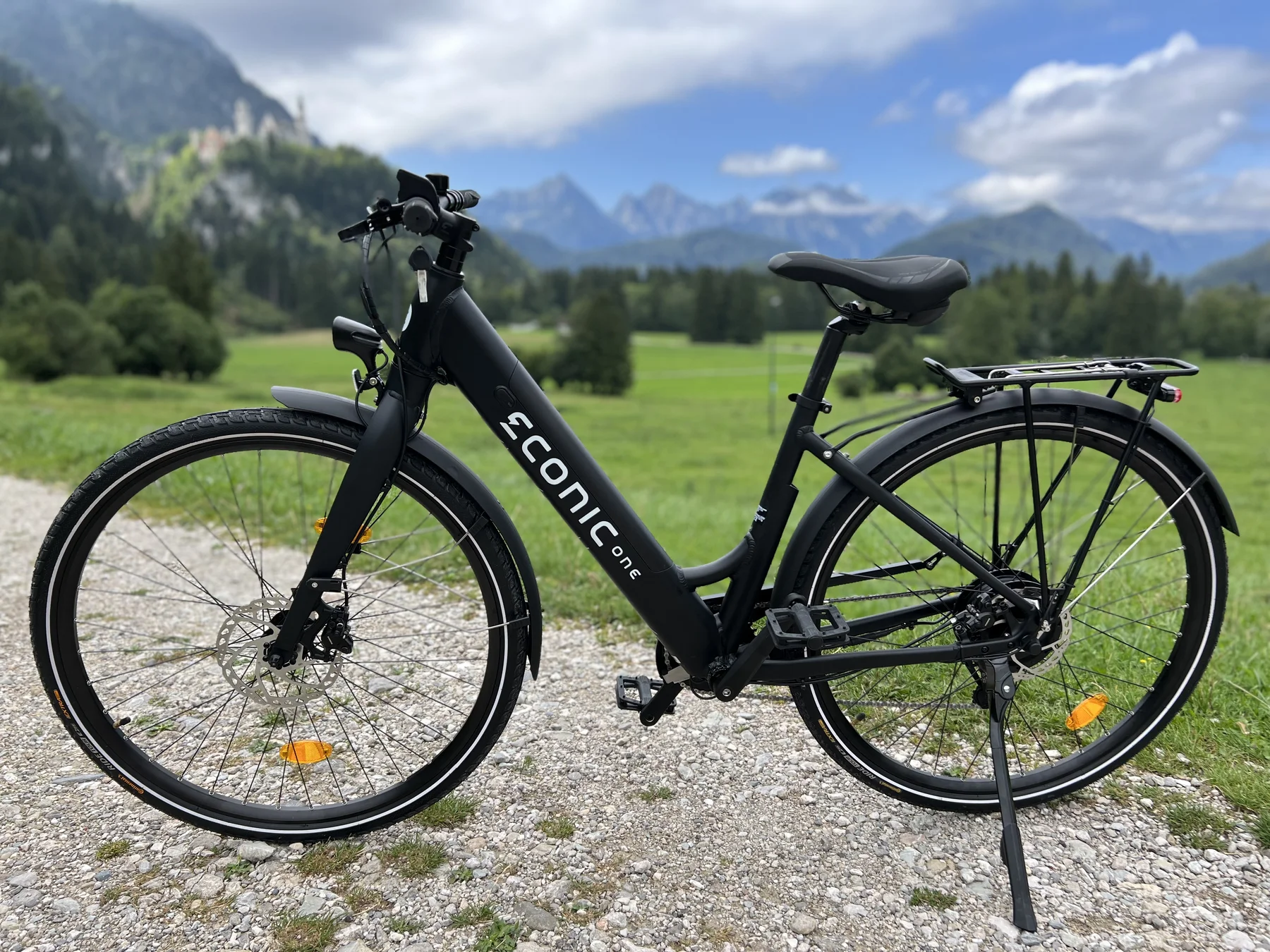 Eonic One Comfort Limited kaufen: Matt-schwarzes E-Bike auf einem Schotterweg vor einem beeindruckenden Alpen-Panorama mit grünen Wiesen und Bergen.