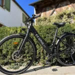 Eonic One Comfort Limited kaufen: Matt-schwarzes City-E-Bike vor einer grünen Hecke und einem ländlichen Gebäude mit Holzschindeln.