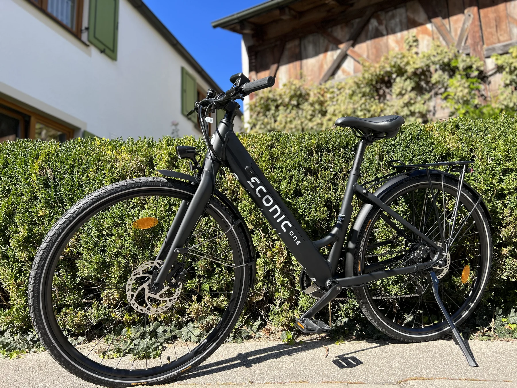 Eonic One Comfort Limited kaufen: Matt-schwarzes City-E-Bike vor einer grünen Hecke und einem ländlichen Gebäude mit Holzschindeln.