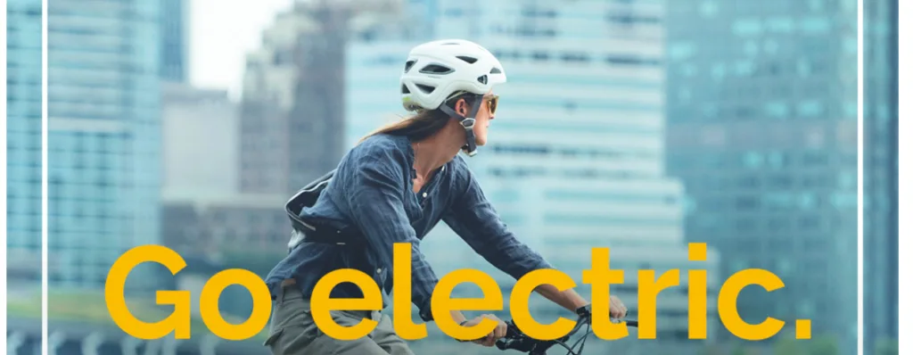 Fahrradfahrerin auf einem E-Bike, Pedelec oder S-Pedelec vor modernen Gebäuden – Schriftzug „Go Electric“ im Vordergrund