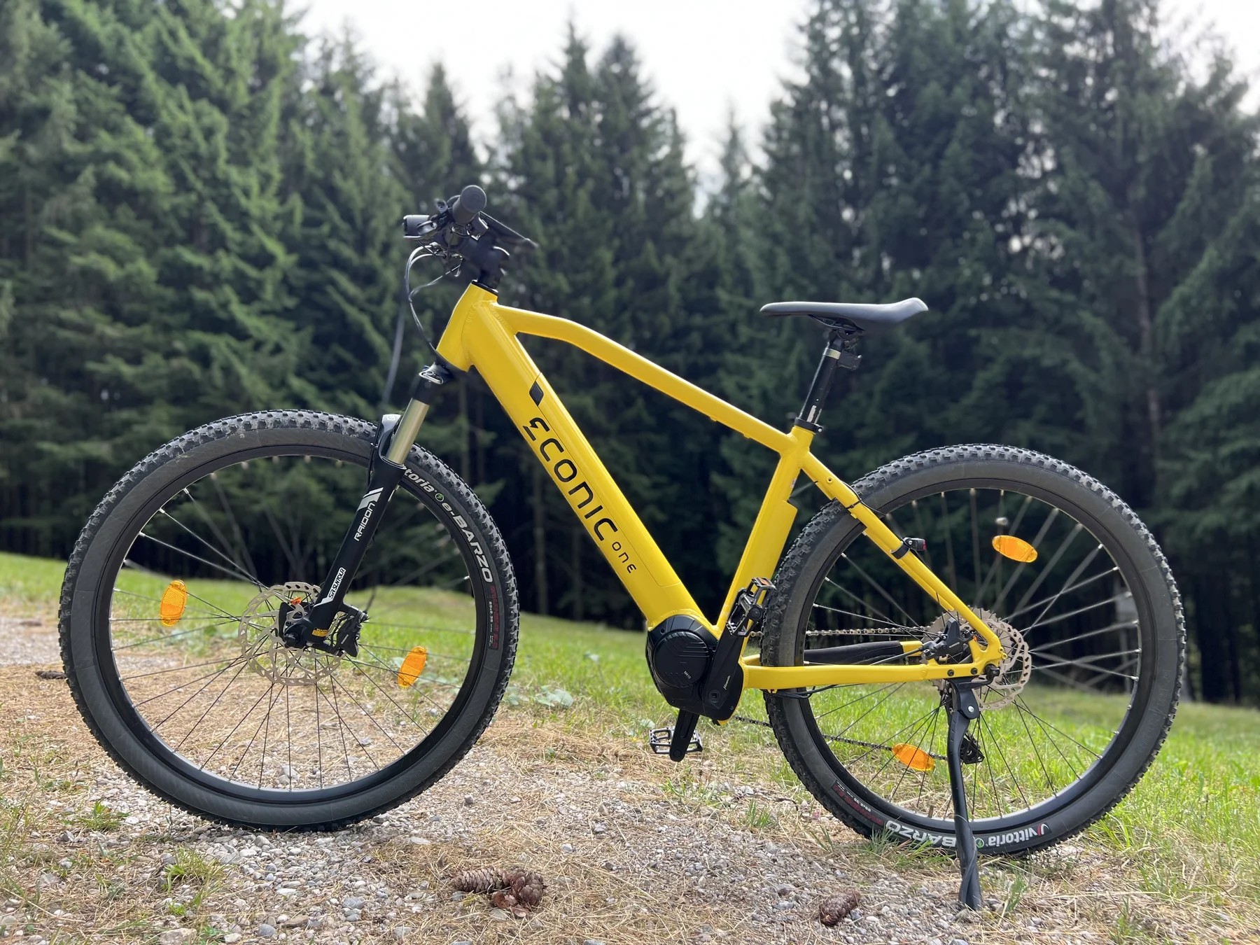 Eonic One Adventure kaufen: Gelbes E-Mountainbike auf einem Schotterweg vor einer Kulisse aus grünen Wiesen und dichten Nadelbäumen.