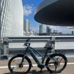 TOTEM Explorer kaufen: Jeansblaues City-E-Bike mit tiefem Einstieg auf Asphalt vor einer modernen Skyline und futuristischer Architektur.