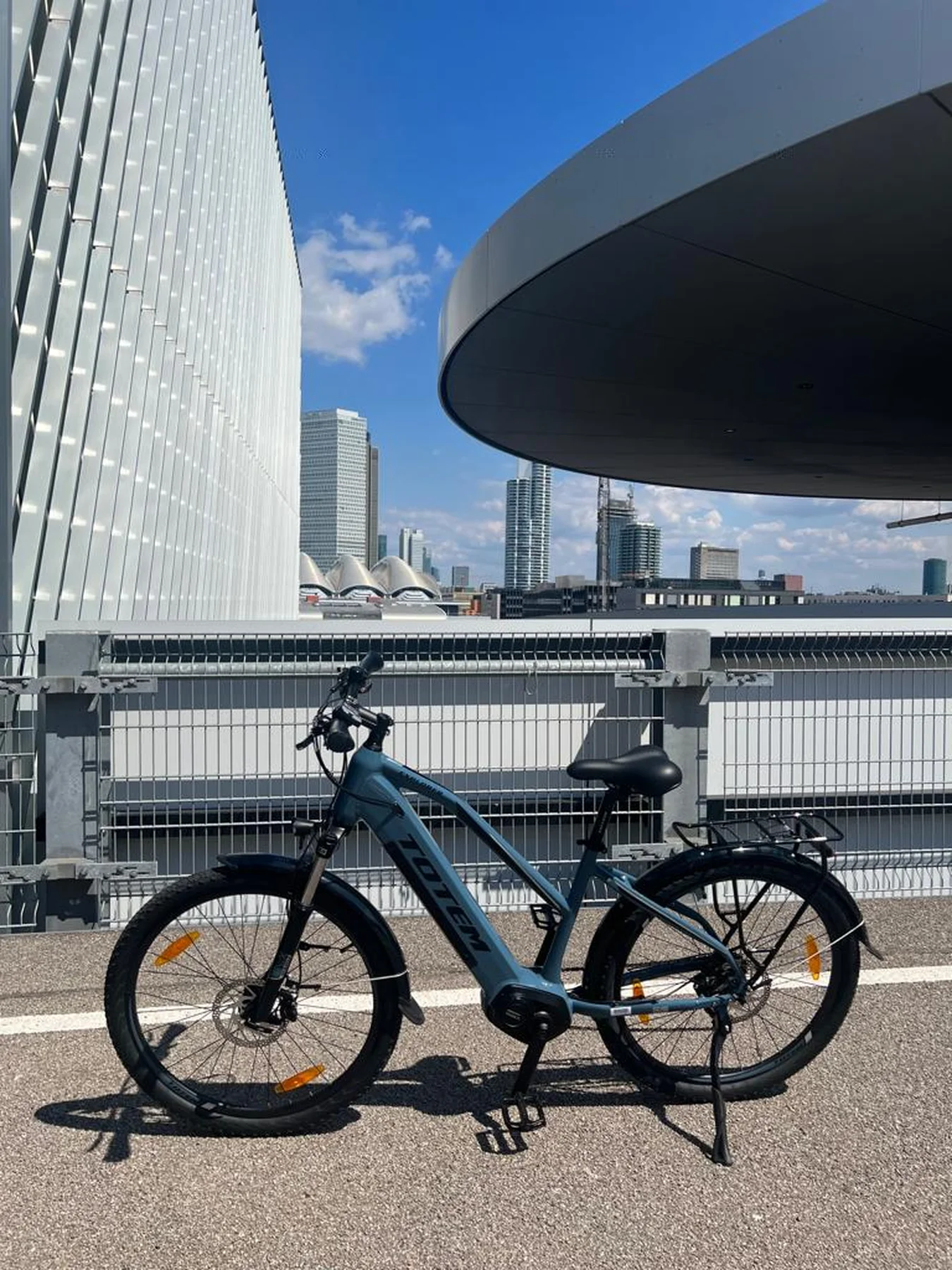 TOTEM Explorer kaufen: Jeansblaues City-E-Bike mit tiefem Einstieg auf Asphalt vor einer modernen Skyline und futuristischer Architektur.