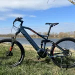 TOTEM Carry kaufen: Blaues vollgefedertes E-Mountainbike auf einer grünen Wiese an einem breiten Flussufer unter blauem Himmel.