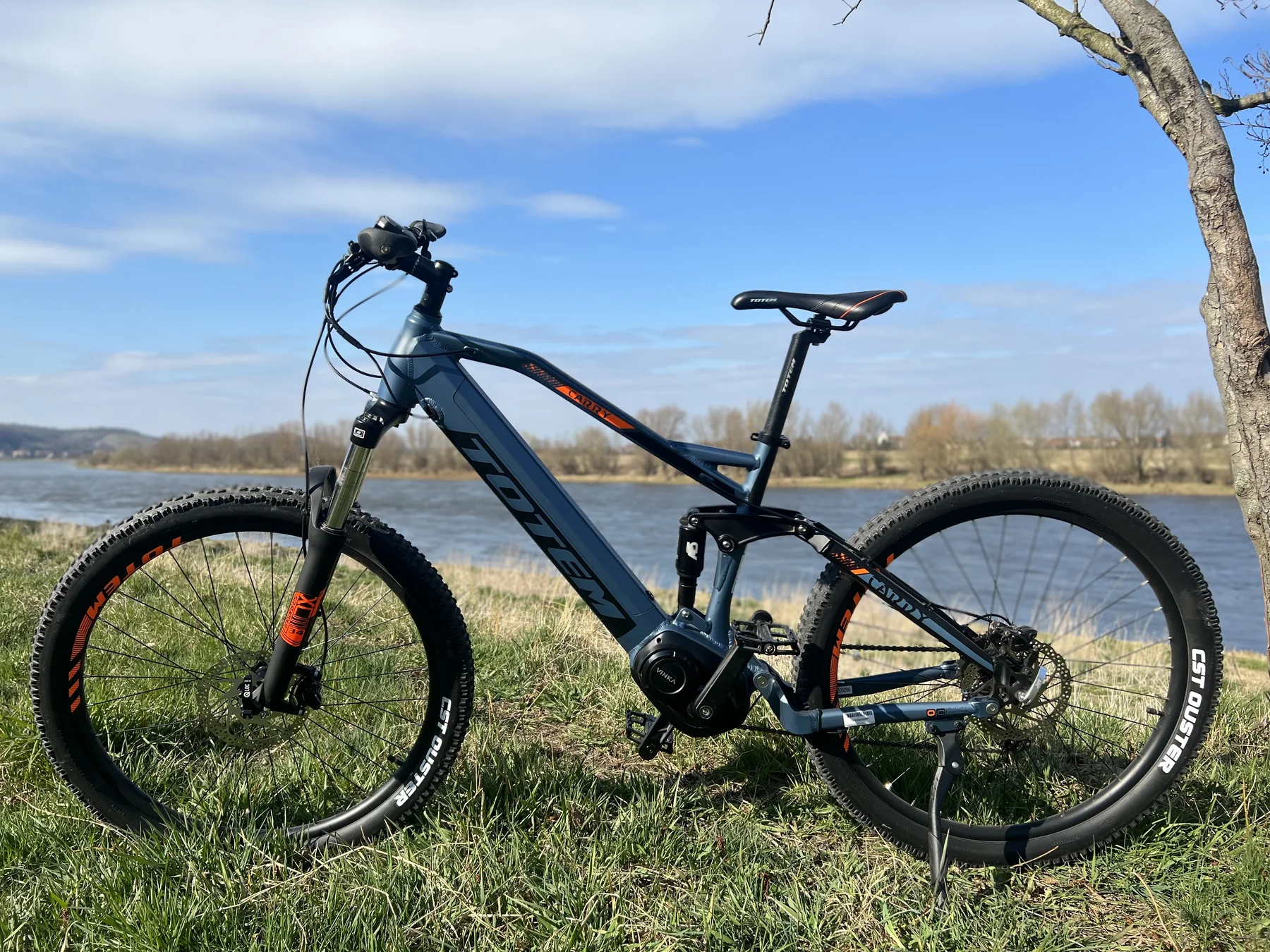 TOTEM Carry kaufen: Blaues vollgefedertes E-Mountainbike auf einer grünen Wiese an einem breiten Flussufer unter blauem Himmel.