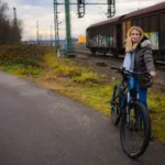 TOTEM Carry kaufen: Junge Frau in Winterjacke mit blauem E-Mountainbike auf einem Asphaltweg neben Bahngleisen und einem Güterzug.