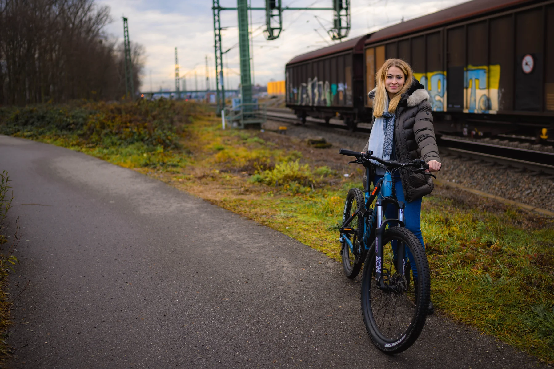TOTEM Carry kaufen: Junge Frau in Winterjacke mit blauem E-Mountainbike auf einem Asphaltweg neben Bahngleisen und einem Güterzug.