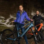 TOTEM Carry kaufen: Ein Paar mit einem blau-grauen und einem orangefarbenen E-Mountainbike vor einer Steinwand mit Graffiti im urbanen Stil.