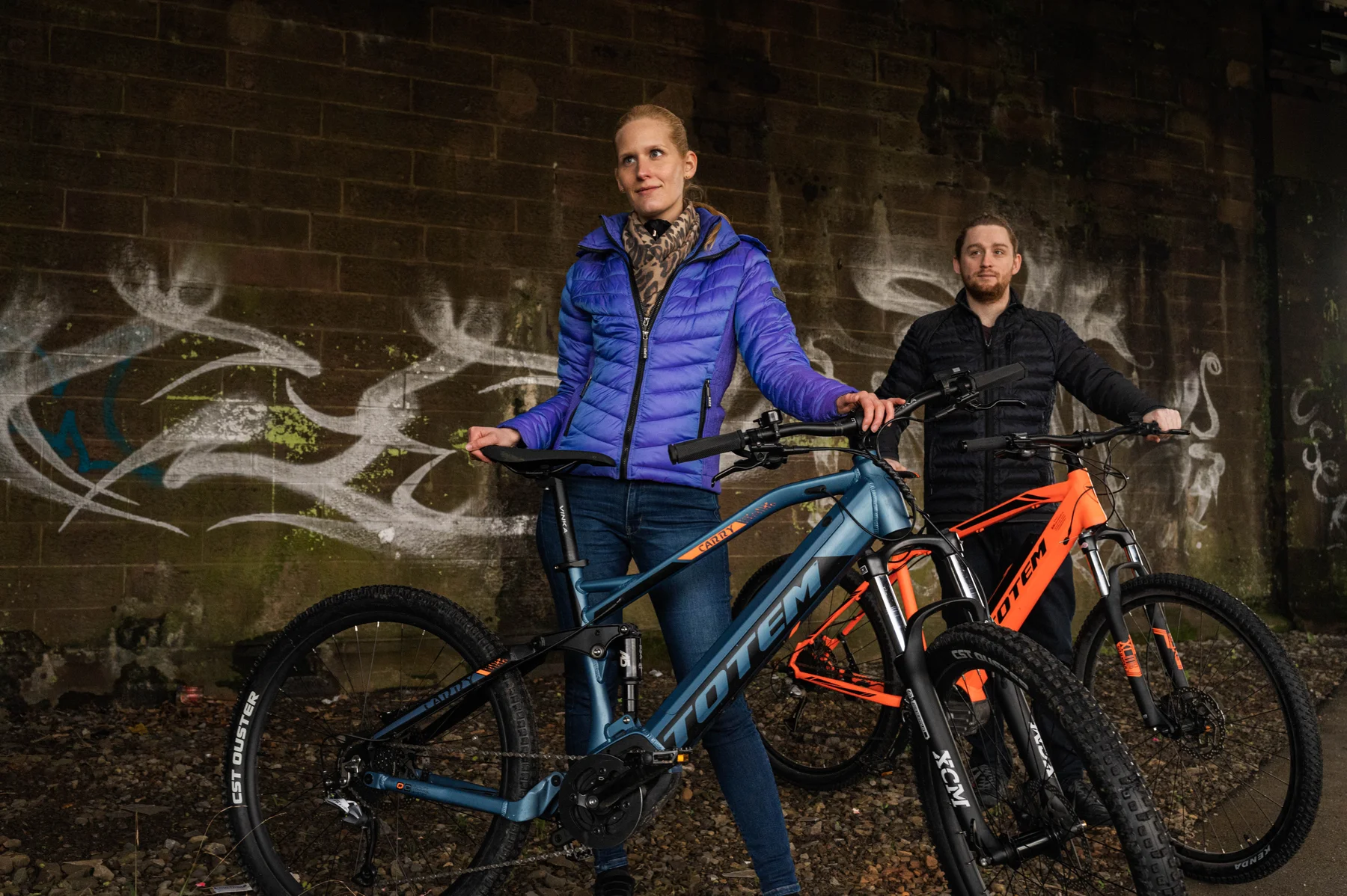 TOTEM Carry kaufen: Ein Paar mit einem blau-grauen und einem orangefarbenen E-Mountainbike vor einer Steinwand mit Graffiti im urbanen Stil.