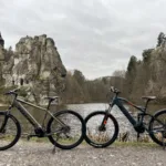 TOTEM Carry kaufen: Ein blaues vollgefedertes E-Mountainbike neben einem grauen Modell an einem See vor der berühmten Felsformation der Externsteine.