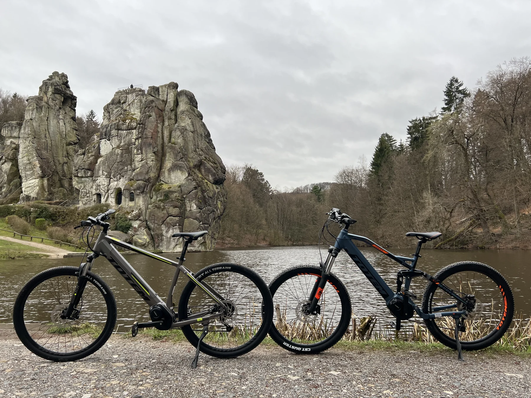 TOTEM Carry kaufen: Ein blaues vollgefedertes E-Mountainbike neben einem grauen Modell an einem See vor der berühmten Felsformation der Externsteine.