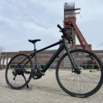 TOTEM Hemner kaufen: Matt-schwarzes Urban-E-Bike auf einem Asphaltplatz vor dem historischen roten Fördergerüst.