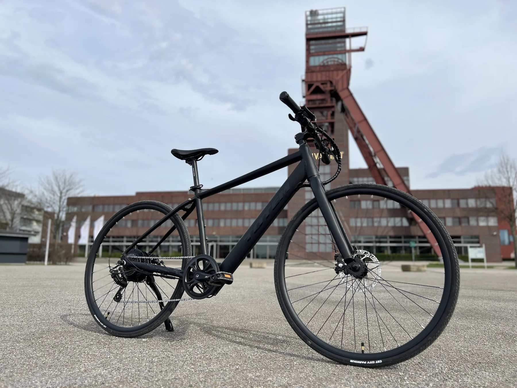 TOTEM Hemner kaufen: Matt-schwarzes Urban-E-Bike auf einem Asphaltplatz vor dem historischen roten Fördergerüst.