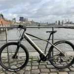 TOTEM Maurice kaufen: Dunkelgraues E-Hardtail-Mountainbike auf Kopfsteinpflaster an einer Uferpromenade vor einer historischen Hafen-Kulisse.