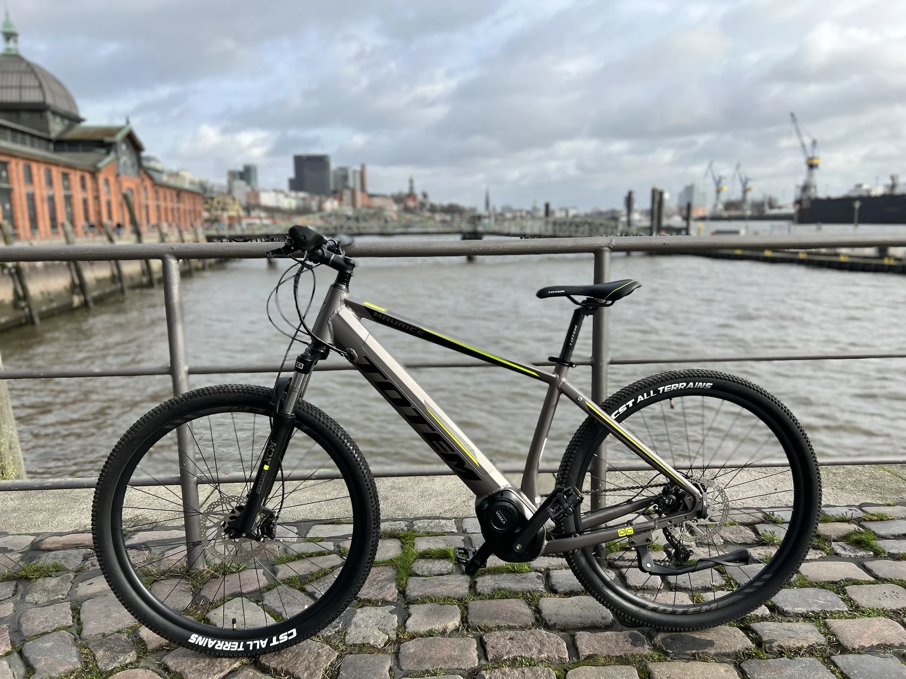 TOTEM Maurice kaufen: Dunkelgraues E-Hardtail-Mountainbike auf Kopfsteinpflaster an einer Uferpromenade vor einer historischen Hafen-Kulisse.