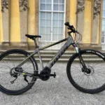 TOTEM Maurice kaufen: Dunkelgraues E-Hardtail-Mountainbike auf einem Kiesweg vor einer barocken Schlossfassade mit großen Sprossenfenstern.