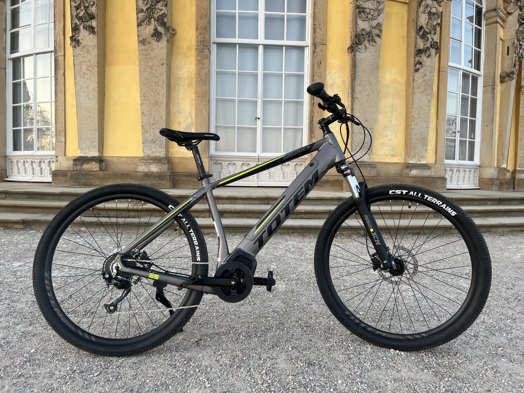 TOTEM Maurice kaufen: Dunkelgraues E-Hardtail-Mountainbike auf einem Kiesweg vor einer barocken Schlossfassade mit großen Sprossenfenstern.