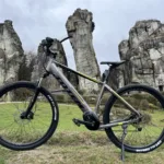 TOTEM Maurice kaufen: Dunkelgraues E-Hardtail-Mountainbike auf einer Wiese vor der imposanten Felsformation der Externsteine im Wald.