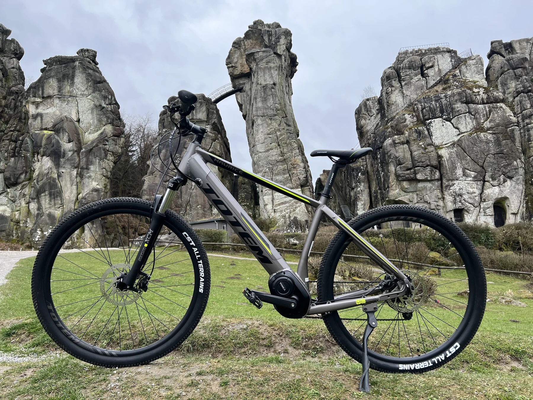 TOTEM Maurice kaufen: Dunkelgraues E-Hardtail-Mountainbike auf einer Wiese vor der imposanten Felsformation der Externsteine im Wald.