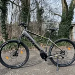 TOTEM Maurice kaufen: Dunkelgraues E-Hardtail-Mountainbike auf einem Schotterweg vor efeubewachsenen Bäumen und einer Betonbrücke.