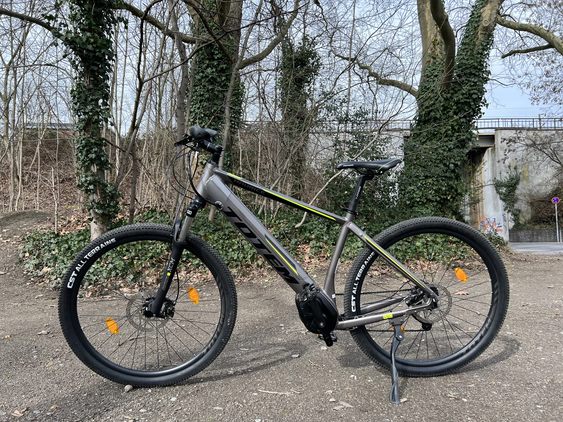 TOTEM Maurice kaufen: Dunkelgraues E-Hardtail-Mountainbike auf einem Schotterweg vor efeubewachsenen Bäumen und einer Betonbrücke.