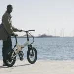 MATE FUSION kaufen: Beigefarbenes Faltrad E-Bike mit dicken Reifen auf einem Holzsteg direkt am Meer mit Windrädern im Hintergrund.