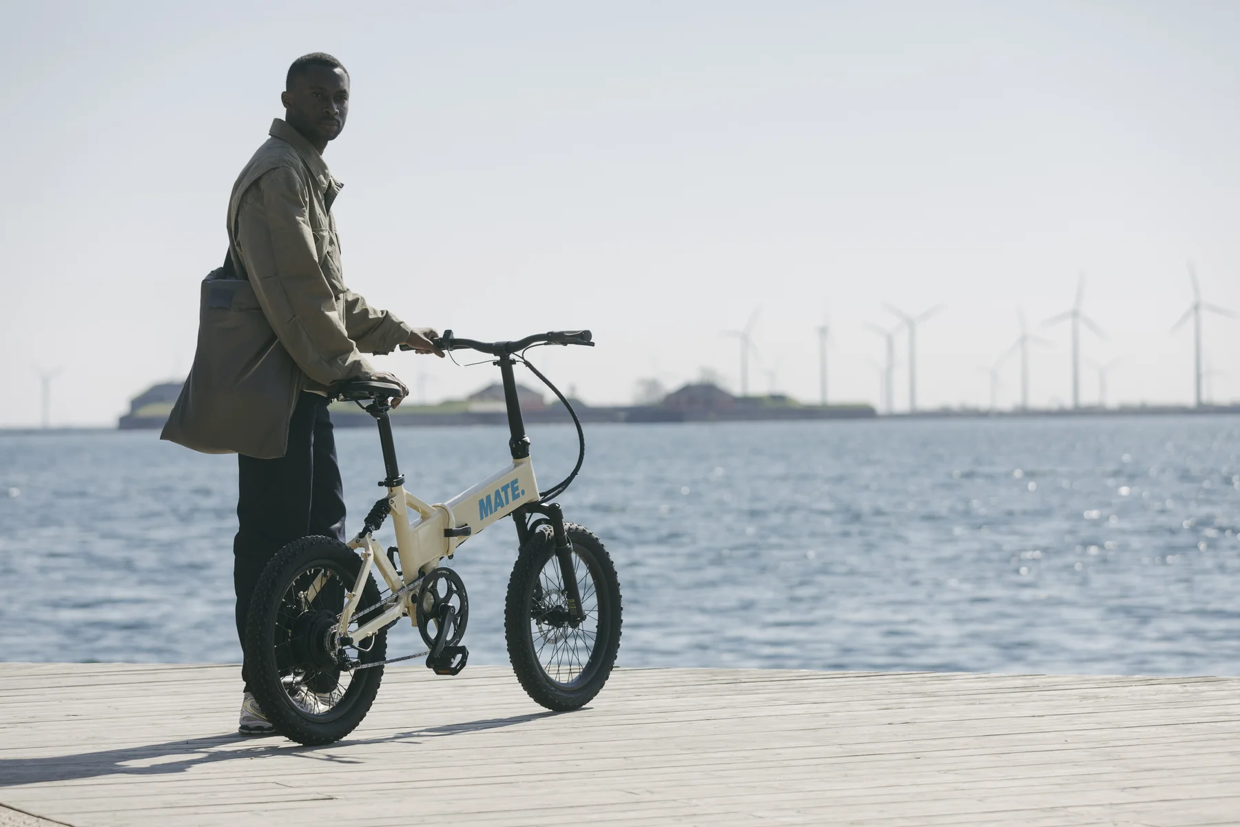 MATE FUSION kaufen: Beigefarbenes Faltrad E-Bike mit dicken Reifen auf einem Holzsteg direkt am Meer mit Windrädern im Hintergrund.