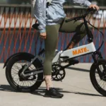 MATE FUSION kaufen: Weißes Faltrad E-Bike mit einer Person in sportlicher Kleidung und olivgrünen Leggings an einer Uferpromenade.