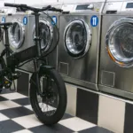 MATE X EVO kaufen: Komplett matt-schwarzes E-Bike auf einem schachbrettartig gefliesten Boden vor einer Reihe großer Industriewaschmaschinen in einem Waschsalon.
