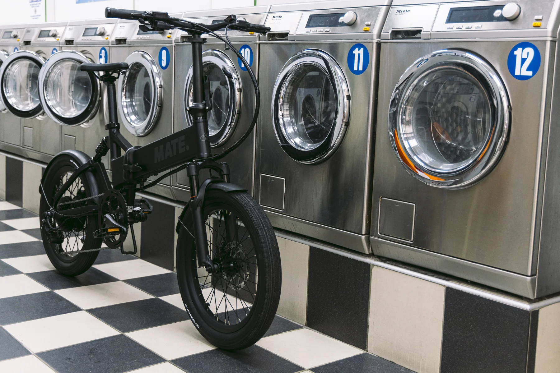 MATE X EVO kaufen: Komplett matt-schwarzes E-Bike auf einem schachbrettartig gefliesten Boden vor einer Reihe großer Industriewaschmaschinen in einem Waschsalon.