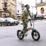 MATE X EVO kaufen: Hellgraues Fatbike E-Bike im echten Fahrbetrieb mit einem stylischen Fahrer auf einer belebten Großstadt-Kreuzung.