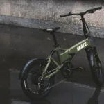 MATE FUSION kaufen: Olivgrünes Faltrad E-Bike auf nassem, spiegelndem Asphalt nach einem Regenschauer in der Stadt.