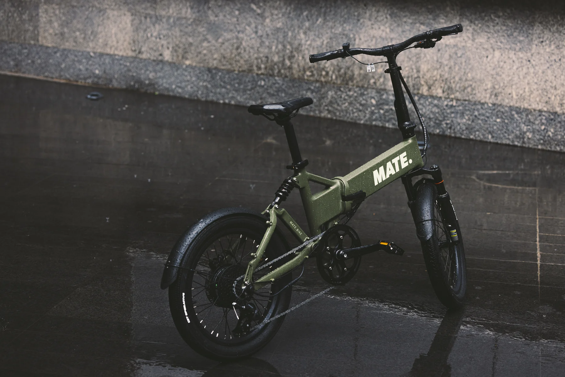 MATE FUSION kaufen: Olivgrünes Faltrad E-Bike auf nassem, spiegelndem Asphalt nach einem Regenschauer in der Stadt.