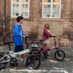 MATE ICON kaufen: Ein junges Paar schiebt ein mintgrünes und ein rosa Faltrad E-Bike gut gelaunt über eine Kopfsteinpflasterstraße vor einer alten Backsteinwand.